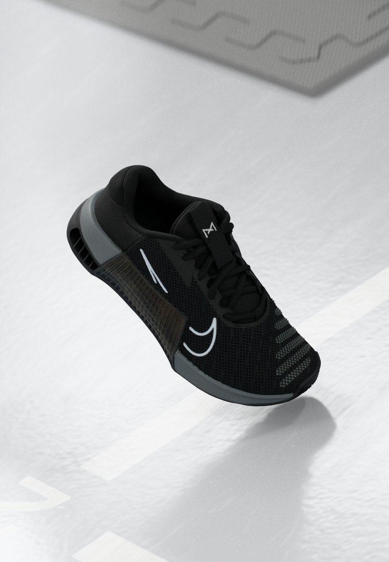 Chaussure de sport noire avec empeigne en mesh, accents gris et semelle intermédiaire à motifs. Présente un logo blanc proéminent et un système de laçage.