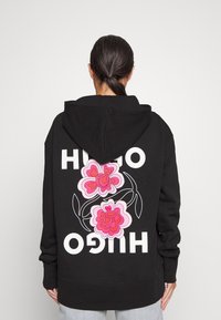 Osoba s tmavými vlasy v culíku, oblečená v černé mikině s růžovým květinovým vzorem a bílým nápisem "HUGO" zrcadleným na zadní straně.