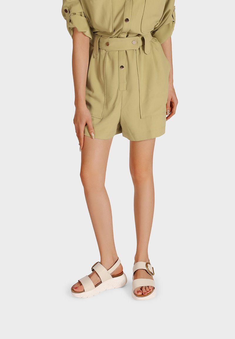 Salopette kaki con maniche corte, vita con cintura, due tasche frontali; abbinata a sandali crema con suole spesse e una fibbia decorativa.