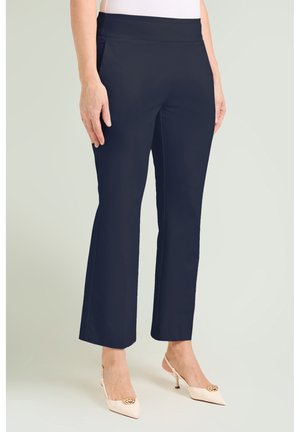 Kobieta ubrana w czarne spodnie 3/4 z bocznymi kieszeniami i beżowe buty na obcasie typu slingback ze złotymi ozdobami, stojąca na jednolitym tle.