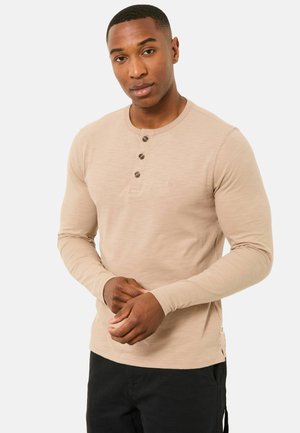 Beiges Henley-Shirt mit langen Ärmeln aus weichem Stoff, mit drei dunklen Knöpfen und einem kleinen gestickten Logo auf der Brust.