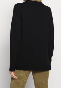 Femme portant un pull noir à manches longues et un pantalon marron texturé, vue de dos sur un fond uni.