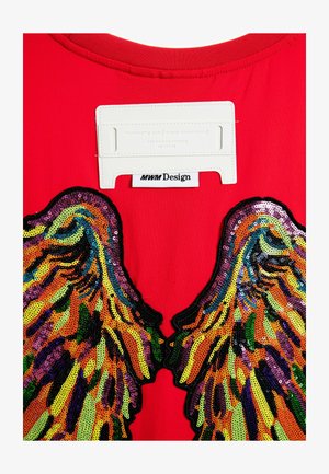 Camiseta roja con grandes apliques de alas de lentejuelas en múltiples colores, con una etiqueta blanca con la inscripción "MWM Design" en la parte de atrás.