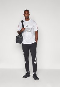Balts Adidas sporta krekls ar melnu logotipu, kombinēts ar tumši pelēkiem biksēm ar baltām sānu svītrām un melniem sporta apaviem. Melna soma.