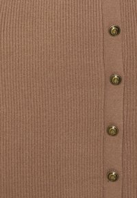 Cardigan marron côtelé avec des boutons dorés sur le côté. Le tissu présente un léger brillant qui apporte texture et profondeur.