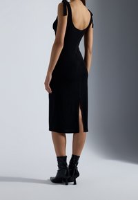 Robe ajustée noire avec dos ouvert et nœuds sur les épaules. Coupe au genou avec fente à l'ourlet. Portée avec des bottines noires.