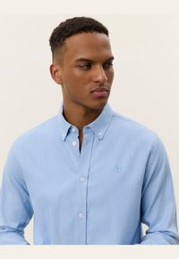 Light blue button-up shirt made of smooth fabric. Features a classic collar, long sleeves, and a small embroidered logo on the chest.