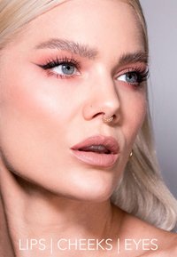 Gros plan d'un visage avec une peau claire, de longs cheveux blonds, un maquillage naturel, un anneau de septum et des détails visibles sur les lèvres, les joues et les yeux.