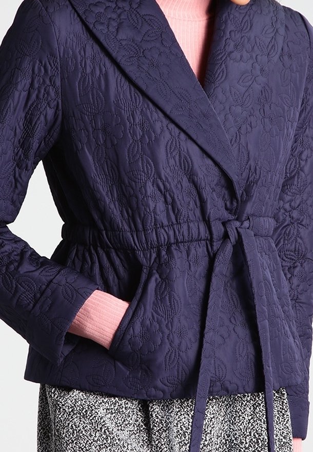 Veste matelassée bleu marine avec surpiqûres florales, ceinture à la taille et poches latérales, portée sur un col roulé rose côtelé et une jupe à motifs noir et blanc.