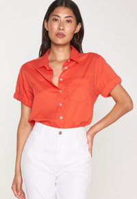 Camicia a maniche corte con taglio morbido di un vivace arancione, dotata di un colletto strutturato, chiusura con bottoni frontale e una singola tasca sul petto, realizzata in tessuto leggero.