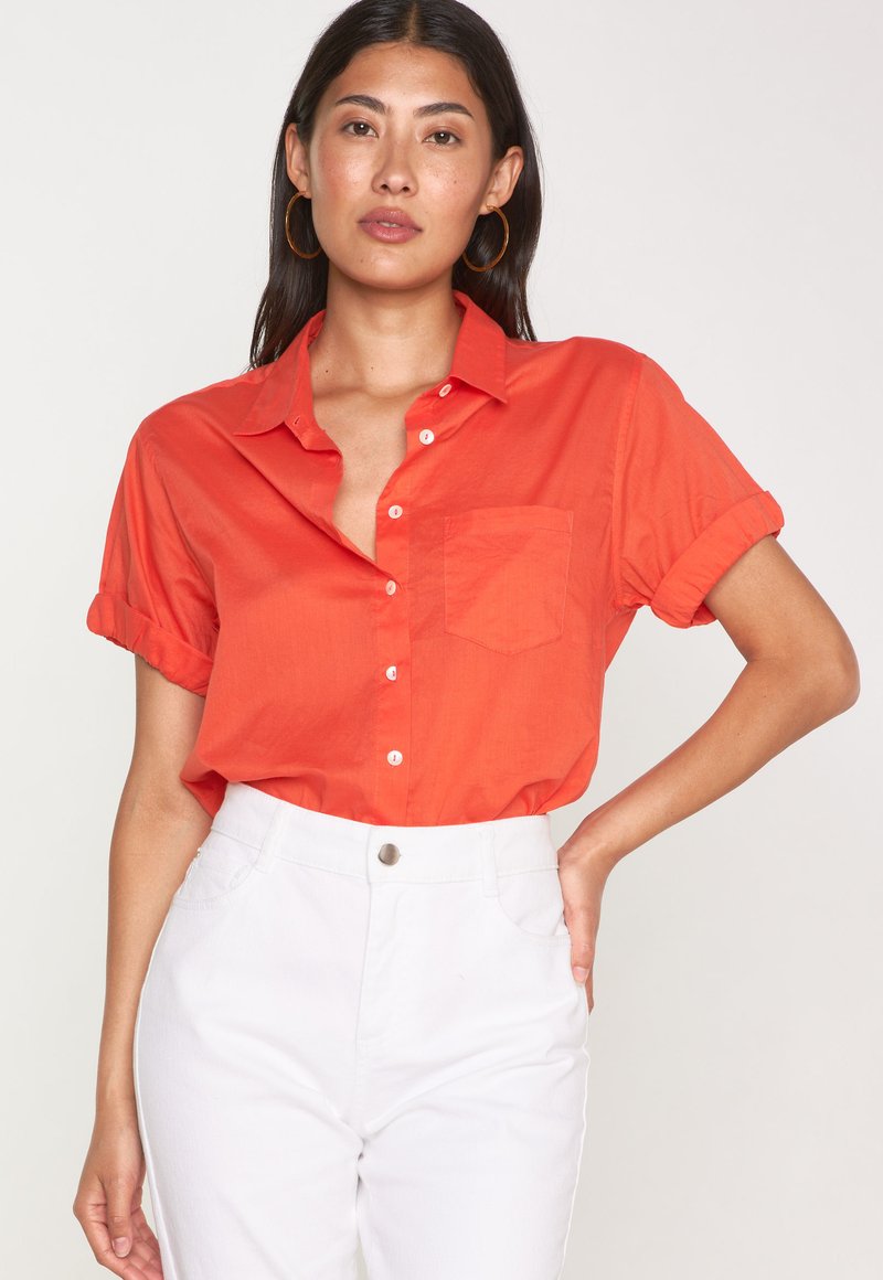 Camicia a maniche corte con taglio morbido di un vivace arancione, dotata di un colletto strutturato, chiusura con bottoni frontale e una singola tasca sul petto, realizzata in tessuto leggero.