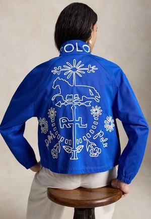 Γυναίκα που φοράει μπλε μπουφάν με λευκή κεντημένη εικόνα αλόγου, ανεμοδείκτη, λουλουδιών και το κείμενο «Polo» στην πλάτη, καθισμένη σε ξύλινο σκαμνί.