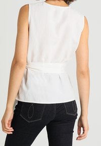Top bianco senza maniche con vita aderente, realizzato in tessuto leggero. Presenta una chiusura con lacci sul retro e una texture liscia. Abbinato a jeans scuri.