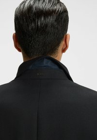 Giacca nera su misura con tessuto testurizzato, caratterizzata da un colletto interno blu in velluto e un dettaglio con logo sul retro.