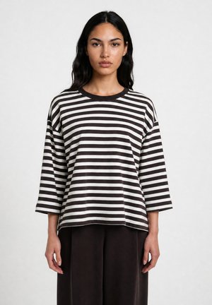 Woman with long dark hair wearing a black and white horizontal striped long-sleeve shirt and dark pants standing against a plain background.
