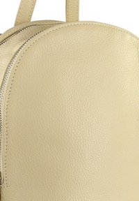 Sac à dos en cuir beige avec une fermeture éclair et des détails de couture visibles, montrant une surface texturée et une partie de deux bretelles.