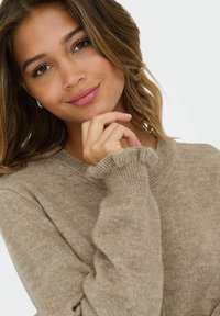 Bežs adīts džemperis ar apaļu kakla izgriezumu, ribotiem piedurknēm un mīkstu tekstūru. Modelis ar viļņainiem matiem tur rokā savu zodu, pārdomājot.