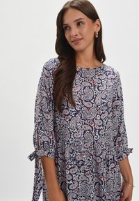Granatowa tunika z nadrukiem paisley w białym, czerwonym i niebieskim kolorze. Posiada rękawy 3/4 z detalami w postaci wiązań. Wykonana z miękkiego materiału, ma luźny krój.