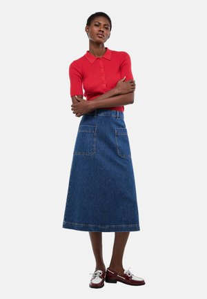 Femme debout, bras croisés, portant un haut rouge à manches courtes et boutons, une jupe en jean bleue mi-longue avec poches avant, et des mocassins bicolores.