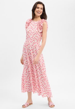 Lächelnde Frau in einem rosa, mit Blumen bedruckten, ärmellosen Kleid mit Rüschen an den Schultern und rosa Slip-On-Sandalen, stehend vor einem weißen Hintergrund.