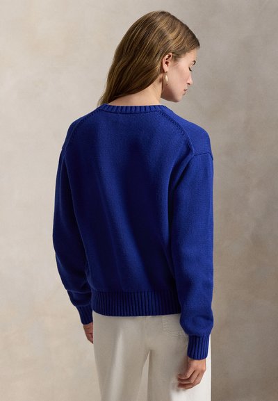 Μπλε πλεκτό πουλόβερ με ριμπ λαιμόκοψη και μανσέτες, με χαμηλωμένους ώμους και χαλαρή γραμμή. Η πίσω όψη δείχνει υφή στο ύφασμα και απλότητα.