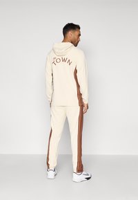 Kreminė džemperis su kelnėmis, derančiomis su rudomis šoninėmis juostelėmis. Ant nugaros yra užrašas "The TOWN". Įeina su jūrine raišteliu ir elastingomis rankovėmis.