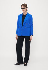 Světlomodrý dvouřadový blazer se zlatými knoflíky, doplněný černým topem, černými širokými kalhotami a černými plochými botami.