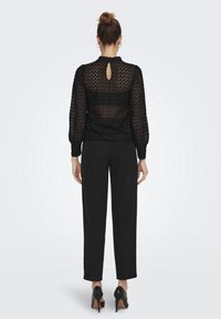 Blusa negra de encaje con detalle de escote en la espalda, mangas largas abullonadas y diseño texturizado, combinada con pantalones negros y tacones.