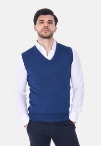 Gilet a maglia blu con scollo a V, indossato sopra una camicia bianca con colletto. Presenta una superficie liscia e orlo a coste. Vestibilità casual.