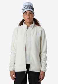Jachetă albă din fleece cu guler în picioare, având un logo pe piept, asociată cu o căciulă alb-negru. Texturată și cu fermoar.