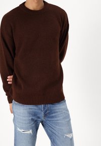 Hnědý pletený svetr s kulatým žebrovaným výstřihem a dlouhými rukávy, spojený s modrými roztrhanými džínami s trhlinami na kolenou.