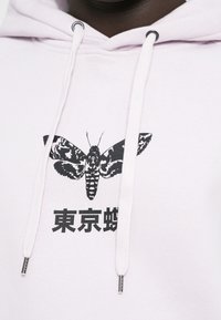 Vit hoodie med svart sabelmålning och japansk text under designen, utrustad med snören med metallspetsar nära halsen.