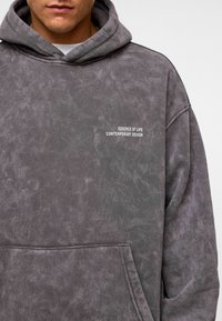 Grijze hoodie met een textuurafwerking, voorzien van een kenguruzak en witte tekst met de boodschap "ESSENCE OF LIFE CONTEMPORARY DESIGN" op de borst.