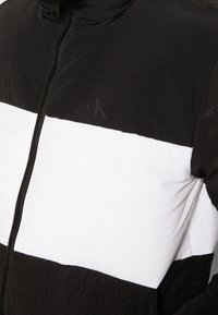 Chaqueta con cierre de color negro y blanco, con bloques de color horizontales y un sutil logo "cK" en el pecho, usada por una persona que mira hacia adelante.