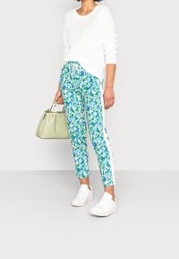 Žena v bílém pleteném svetru, květinových modro-zelených kalhotách, bílých teniskách, držící světle zelenou kabelku na jednoduchém pozadí.