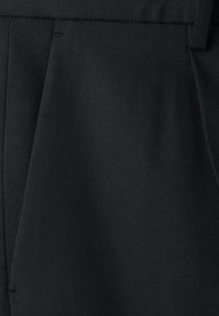 Nahaufnahme von schwarzen, maßgeschneiderten Hosen mit Taillenbund, Vorderfalz und Gürtelschlaufen aus fein strukturiertem Stoff.