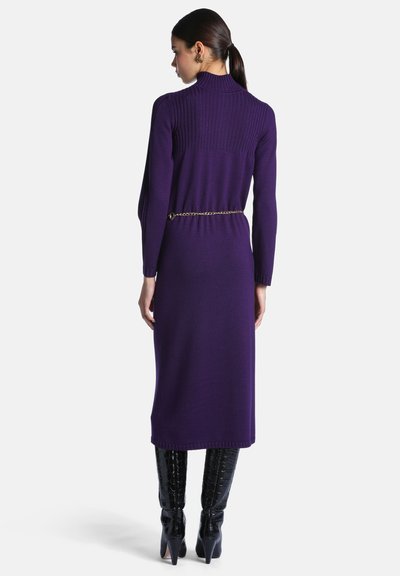 Femme aux cheveux foncés en queue de cheval, debout de dos, portant une longue robe en maille violette avec une ceinture chaîne dorée et des bottes noires montantes à talons.