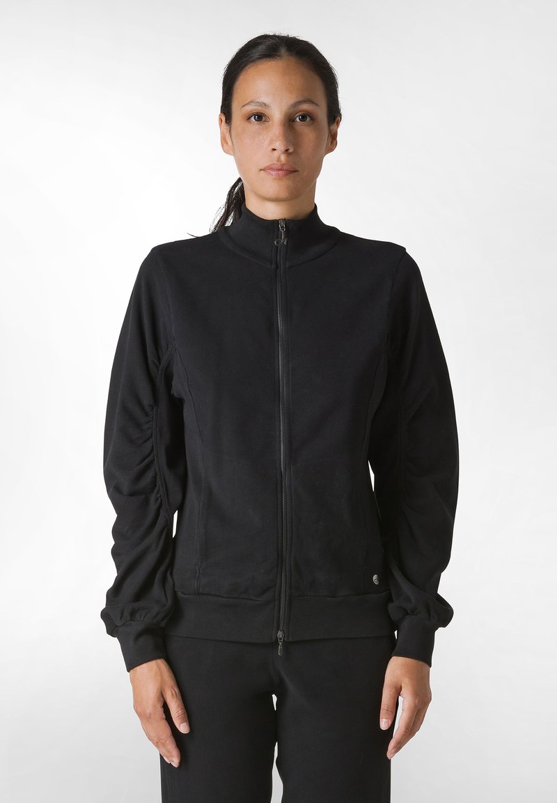 Giacca nera con zip, colletto alto, maniche arricciate e polsini a costine. Realizzata in un materiale morbido, progettata per un look aderente.