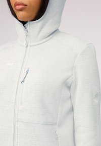 Giacca in pile grigia chiara con chiusura a zip, cappuccio e tessuto testurizzato. Presenta una tasca sul petto e un logo decorativo sulla manica.