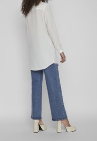 Camicia bianca a maniche lunghe, orlo dritto, abbinata a jeans in denim blu e scarpe con tacco alto color crema con accento nero sul tacco.