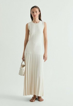Mujer vistiendo un vestido sin mangas de color crema con detalles de tachuelas y falda plisada, sosteniendo un bolso blanco y calzando sandalias con tiras.