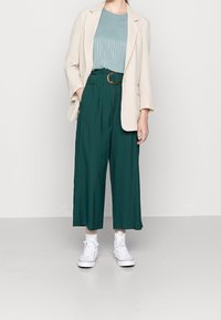 Beżowa kratkowana marynarka na jasnoniebieskiej bluzce, zestawiona z szerokimi, ciemnozielonymi spodniami i białymi trampkami za kostkę.