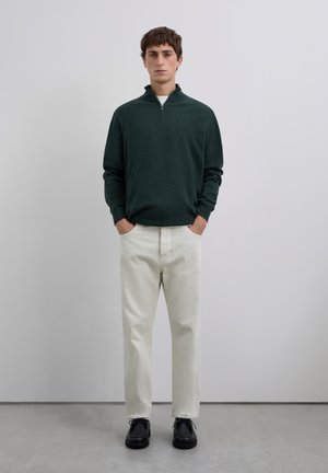 Jeune homme debout, les mains dans les poches, portant un pull vert foncé à fermeture éclair quart, un pantalon blanc cassé et des chaussures noires sur un sol gris.