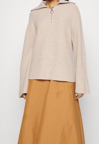 Hellbeiger, gerippter Pullover mit einem halben Zipperkragen und silbernem Ringzug, kombiniert mit einem rostfarbenen ausgestellten Rock. Weiche Textur, oversized Passform.