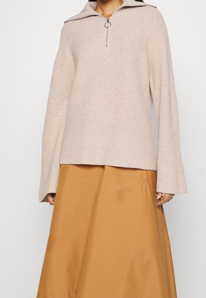 Hellbeiger, gerippter Pullover mit einem halben Zipperkragen und silbernem Ringzug, kombiniert mit einem rostfarbenen ausgestellten Rock. Weiche Textur, oversized Passform.