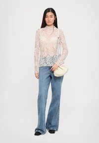 Top de encaje de manga larga en un tono beige claro, con un patrón floral, superpuesto sobre un borde de encaje más oscuro. Combinado con jeans acampanados azules y un bolso de crema.