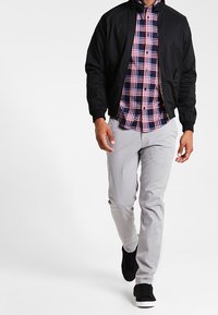 Giubbotto bomber nero sopra una camicia a quadri blu e rossi, pantaloni grigi e sneaker nere. Presenta una vestibilità elegante e moderna con motivi contrastanti.