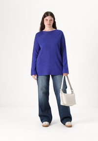 Pulover lila, cu maneci lungi și croi lejer, cu un model texturat, combinat cu blugi evazați de culoare albastru închis și o geantă de umăr bej deschis.