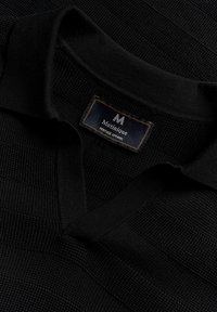 Črna polo majica z zgoščenim pletenim vzorcem. Ima rebrasto ovratnik in oznako z izvezenim napisom "Matinique Heritage Apparel" v kontrastni barvi.