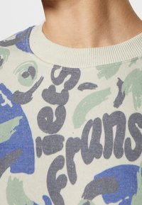 Gros plan sur une personne portant un sweat-shirt beige avec des motifs abstraits bleus et verts, ainsi qu'un texte partiel gris foncé près du col.